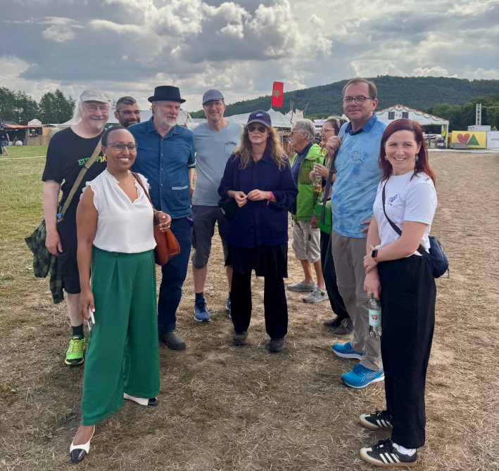 Das Foto zeigt zehn Menschen. Sie stehen in einem lockeren Halbkreis und schauen zum Betrachter. Im Hintergrund sieht man das Festival-Gelände.
