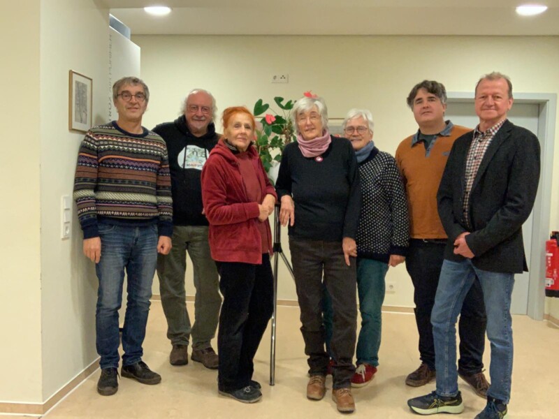 Das Foto zeigt sieben Personen in einem Flur stehend. In der Mitte stehen drei Frauen, links und rechts davon jeweils zwei Männer. 